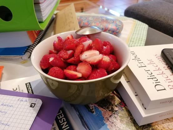 Eine grüne Müslischüssel aus Porzellan ist gefüllt mit vielen geviertelten roten Erdbeeren. Die Schüssel steht auf einem Glastisch, der zu voll ist mit Ordnern, Papier und Büchern.