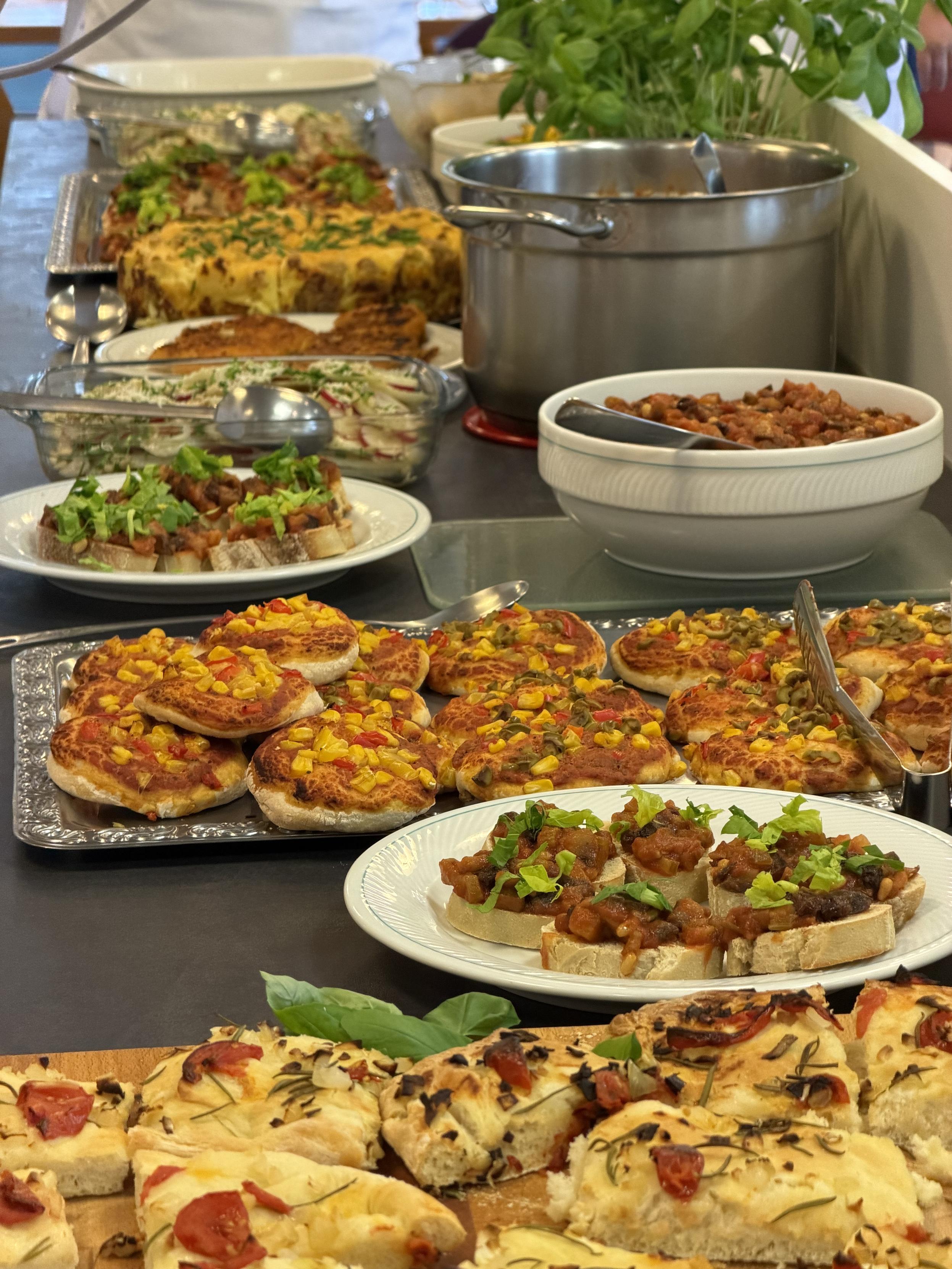 Reich gedecktes Buffet eines veganen Kochkurses mit Mini-Pizzen, Bruschetta, Reisgericht, Aufläufen, frischem Basilikum und weiteren pflanzlichen Speisen.